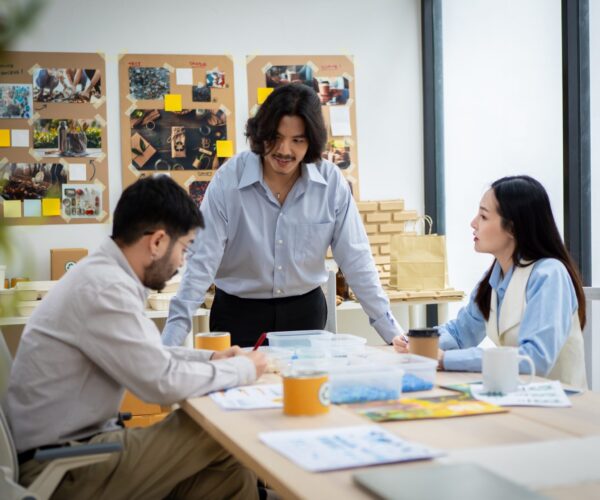 Three people are sitting at a table in a room with a whiteboard on the wall. A woman is writing on a piece of paper with a pen. Scene is collaborative and focused